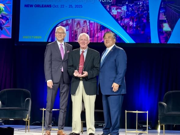 Three men in suits and ties standing on a stage. The man in the middle is holding a clear award trophy.